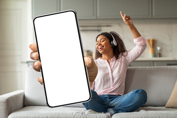 A joyful black woman sits on a couch, listening to music through headphones. She holds a smartphone...