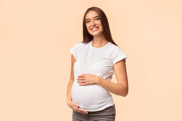Obraz premium Happy Pregnancy Concept. Expectant Mother-To-Be Touching Belly Smiling At Camera Standing Over White Background. Studio Shot