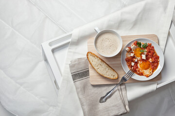 A cozy breakfast tray setup featuring sunny-side-up eggs with tomato sauce and feta cubes, served with a slice of rustic bread. A cup of frothy coffee completes the perfect breakfast in bed.