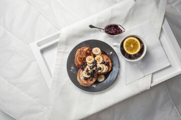 A delightful breakfast tray featuring pancakes topped with banana slices and blueberries, served with a side of berry jam. A cup of tea with a slice of lemon completes the cozy setting.