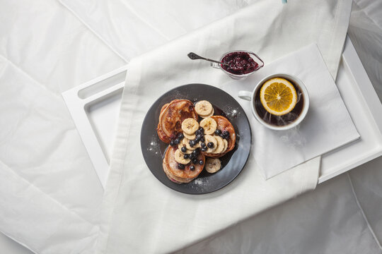 A delightful breakfast tray featuring pancakes topped with banana slices and blueberries, served with a side of berry jam. A cup of tea with a slice of lemon completes the cozy setting.
