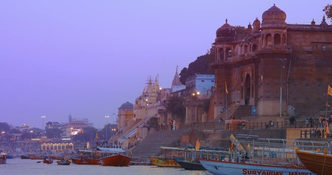 Varanasi, India. Many Boats Moored On Ganga River Near Ghats. General View On Old Red Wall Chet Singh Fort, Jain Ghat, Vaccharaja Ghat. Purple Sunset Sky At Evening Time. Camera Moving Along Riverbank
