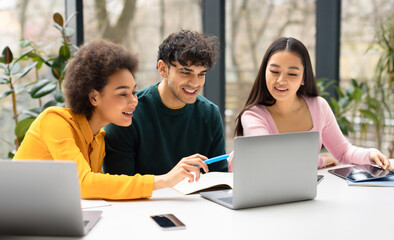 Happy international students friends sitting at desk in coworking space and using laptop, making project together or watching educational video, panorama