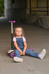 Naklejka premium The little girl is tired of riding her scooter. She's sitting on the ground tired