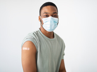 Coronavirus Vaccination. Portrait Of Vaccinated African Guy Wearing Protective Face Mask Looking At Camera Posing With Adhesive Tape On Arm After Vaccine Injection Over White Background