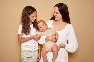 Satisfied adorable female parent with baby and older daughter posing indoor isolated over beige...