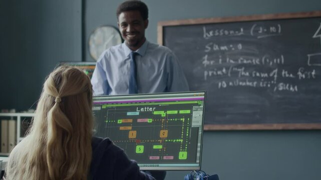 Medium shot of Black male programming teacher speaking to Caucasian school girl sitting at desk in front of computer monitor displaying software interface and raising hand to ask question