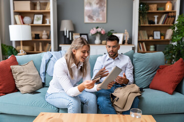Obraz premium Shopaholic concept. Young business couple sitting at home on sofa having argue about woman spending the money, buying and ordering online with husband credit card, using digital tablet for orders.