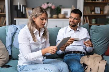 Shopaholic concept. Young business couple sitting at home on sofa having argue about woman spending the money, buying and ordering online with husband credit card, using digital tablet for orders.