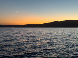 A golden sunset over Bracciano Lake in Trevignano Romano, Italy, with a crescent moon rising above the calm evening waters.