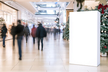 An empty poster mock-up for advertisement in a shopping center; a blank white billboard template surrounded by Christmas conifer decoration and garlands near the stairway down to a mall hall