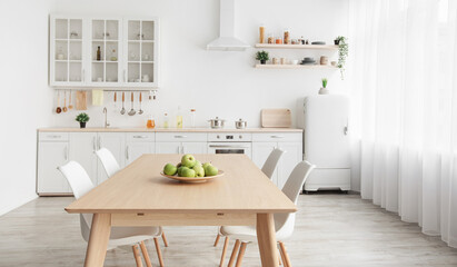 Contemporary minimalist interior of kitchen and dining room. White furniture with utensils and dinner table with chairs in Scandinavian style. Modern light design