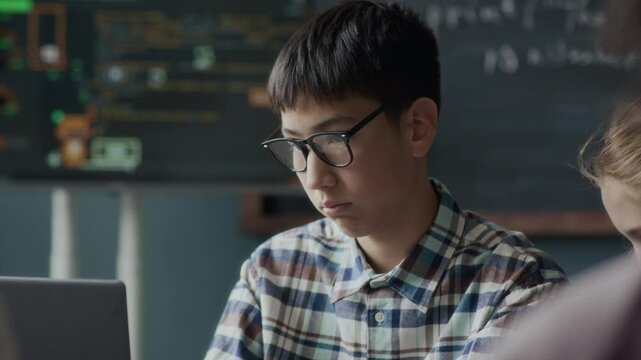 Waist up of Asian teenage schoolboy in glasses and played shirt typing on laptop keyboard while studying coding and programming in class during lesson