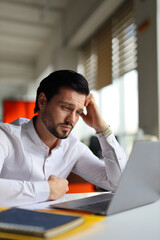 A man is sitting at a desk with a laptop and a notebook. He is looking at the laptop with a frown on his face