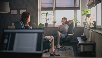 African American teenage girl sits in chair, discusses problems and psychological trauma on counseling therapy session with female psychologist or psychotherapist. Laptop in the foreground. Psychology