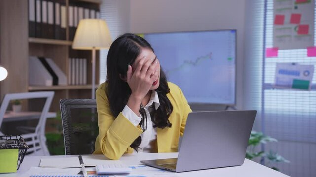 Young asian businesswoman is experiencing stress while working on her laptop in her office. She is massaging her temples and looks concerned