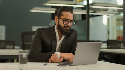 Busy concentrated Indian male employer company corporate Arabian entrepreneur boss CEO business man Muslim businessman at office with laptop working typing thinking solution idea pensive looking away