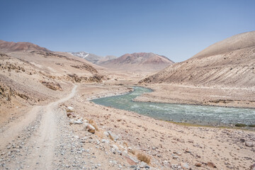 Dusty road of the Pamir Highway winds and twists in the valley of the Tien Shan Mountains in Tajikistan in the Pamirs and the Pamir River with blue clear glacial water flows in the valley