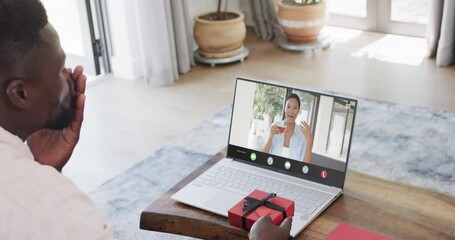 African american man holding red gift and using laptop with biracial woman on screen - Powered by Adobe