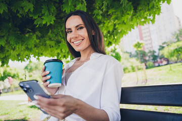 Fototapeta premium Photo of pretty young good mood girl drink coffee hold device sit bench wear white clothes walk outside urban city park
