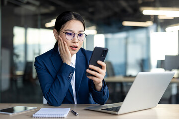 Asian businesswoman shows surprised expression looking at phone in office. Uses laptop and notepad, depicting multitasking and communication tech. Professional attire suggests corporate or business