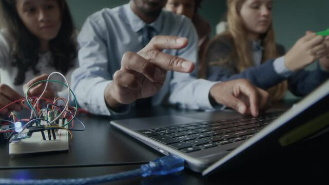 Stab shot of unrecognizable Black male programming teacher typing on laptop keyboard while explaining material to multiethnic group of gen z schoolkids during lesson