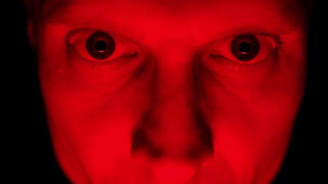 Angry man face under red lighting, showing intense emotions of rage and hostility. Close-up.