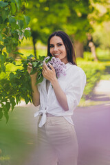 Vertical hoto of pretty young good mood girl posing smell lilac flowers bush wear white clothes walk outside urban city park