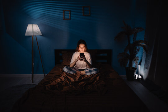 Tired teenage girl browses information on smartphone before sleep on bed in semi-dark bedroom