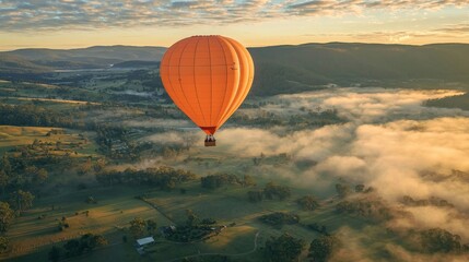 Obraz premium A bright orange hot air balloon floating over a scenic landscape, the morning mist rising from the valleys below