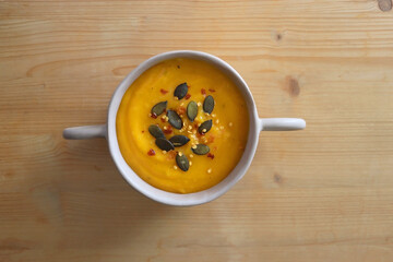 Bowl of creamy pumpkin and potato soup, with pumpkin seeds and chilli flakes toppings. Top view, wooden background.