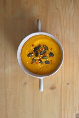 Bowl of creamy pumpkin and potato soup, with pumpkin seeds and chilli flakes toppings. Top view, wooden background.