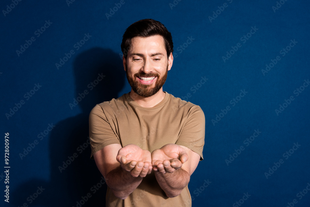 Poster Photo of positive grateful man with beard dressed beige t-shirt palms hold object empty space isolated on dark blue color background