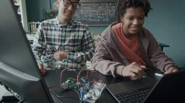 Waist up of two multiethnic young boys sitting in front of laptop at desk with circuit board parts and wires on it and working on electronics school project in classroom