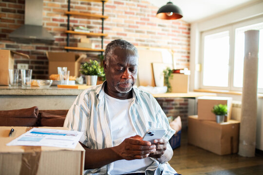 Senior black man using smartphone in home full of moving boxes