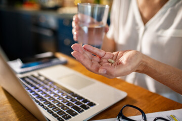 Close up senior woman taking supplements or vitamins while working on laptop at home
