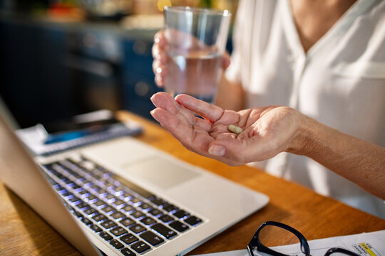 Close up senior woman taking supplements or vitamins while working on laptop at home