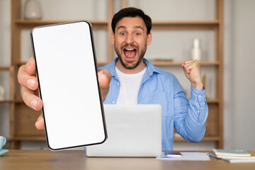 A man sitting at a desk in his home office joyfully raises a smartphone while celebrating a recent...