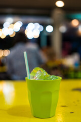 A green plastic cup with ice cube and drink straw on yellow wood table and bokeh background
