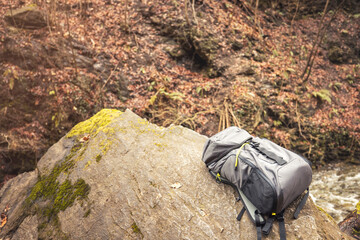 Nestled on a mossy boulder, a well-used backpack awaits by the river's edge, surrounded by the warm hues of autumn leaves and gentle water flows