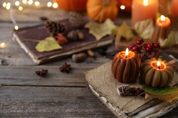 Burning candles in shape of pumpkins, old book and autumn decor on wooden table, closeup
