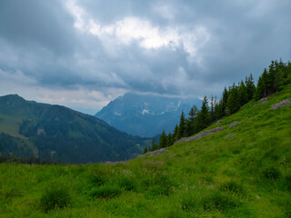 Obraz premium Hiking trail along alpine meadow with panoramic view of cloud covered mountain peaks in Eisenerz Alps, Styria, Austria. Tranquil atmosphere in Austrian Alps in spring. Outdoor trip on overcast day
