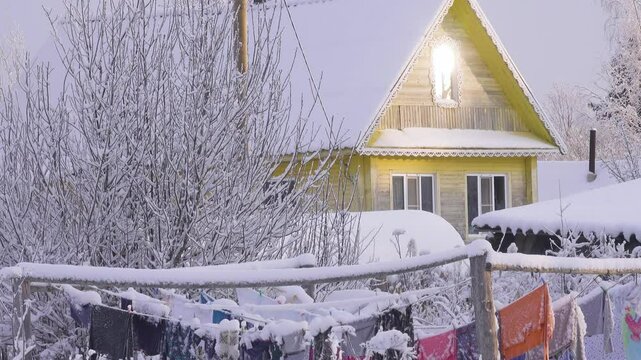 Siberia village in winter, rural idyll life. Wooden snow covered ancestral house, sun shines in attic carved window, snow-covered linen drying on hangers. Sheets and towels smell of frost. Nice view