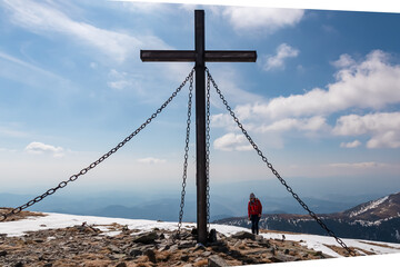 Obraz premium Hiker woman reaching wooden summit cross standing atop mountain peak Lenzmoarkogel, Gleinalpe, Lavanttal Alps, Styria, Austria. Hiking trail on snow covered alpine meadow. Wanderlust. Tranquil serene