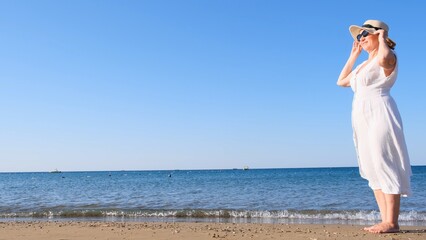 mature woman in a straw hat and white dress enjoying the sun walking along the blue sea coast on a sunny summer day, enjoying freedom and relaxation. The concept of a senior citizen everyday life