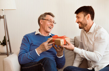 Mature Son Congratulating Elderly Father Giving Him Birthday Gift Sitting On Sofa At Home. Selective Focus