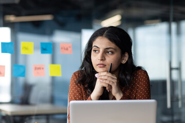 Young thinking business woman close up at workplace inside office, female company worker serious thoughtfully looking out window, working with laptop, company worker focused.