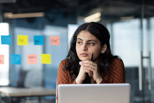 Young thinking business woman close up at workplace inside office, female company worker serious thoughtfully looking out window, working with laptop, company worker focused.