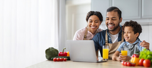 A joyful African American family engages in a cooking session at home, surrounded by fresh vegetables and drinks. They smile as they work on a laptop, planning their meal together.