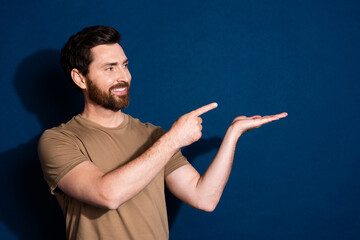 Photo of cheerful man with beard dressed beige t-shirt look directing at product on arm empty space isolated on dark blue color background